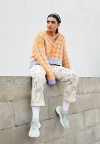 Jeune femme aux cheveux foncés et serre-tête, assise sur un rebord en béton, portant un pull duveteux orange et lavande, un pantalon à motifs et des baskets vertes.