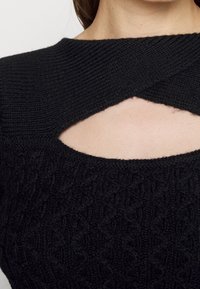 Gros plan sur un pull en maille noire avec un détail entrecroisé et une ouverture en forme de goutte au niveau du haut de la poitrine, porté par une personne.