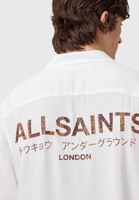 Witte short-sleeve shirt met een grote, tekstuurlijke print van "ALLSAINTS" met een slangenhuidpatroon op de achterkant, en aanvullende tekst in het Japans.
