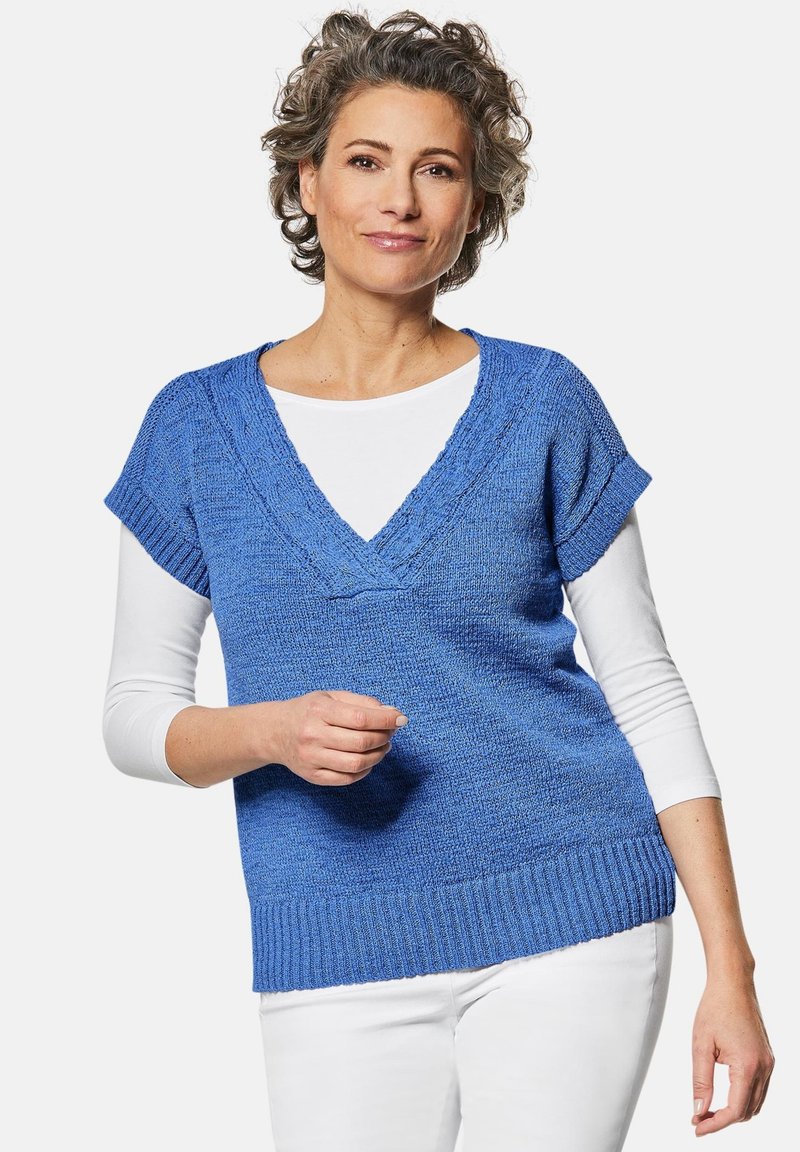 Blauwe gebreide trui met korte mouwen en een V-hals, met gestructureerde stof en ribbelzoom, gelaagd over een witte longsleeve top.