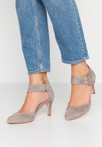 Harmaat mokkanahkakengät korkoilla, teräväkärkiset ja nilkkaraskailla, joissa koristeena solki ja hoikka korko, yhdistettynä lyhytlahkeisiin sinisiin farkkuihin.