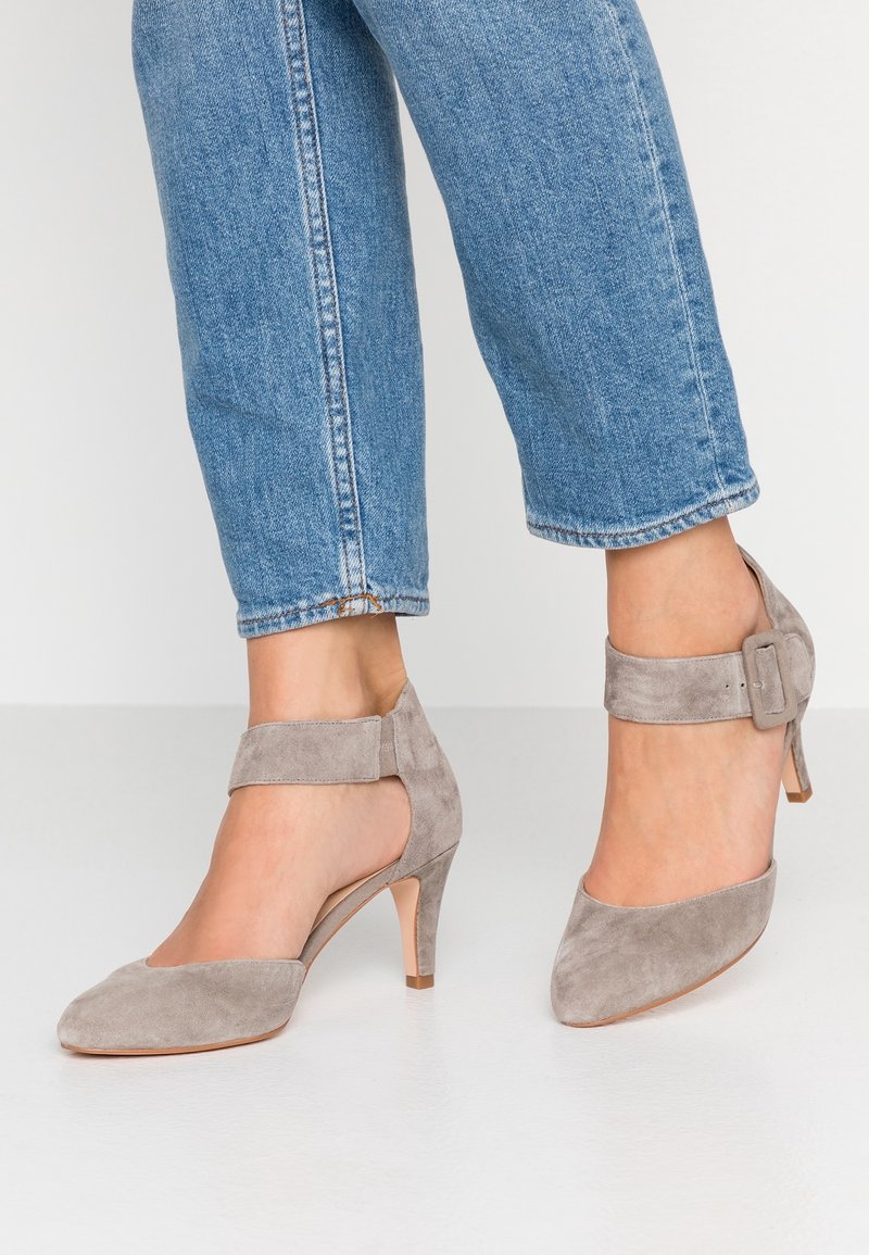 Harmaat mokkanahkakengät korkoilla, teräväkärkiset ja nilkkaraskailla, joissa koristeena solki ja hoikka korko, yhdistettynä lyhytlahkeisiin sinisiin farkkuihin.