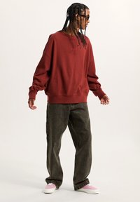 Sudadera oversized en color burdeos con puños acanalados, combinada con jeans holgados de color verde oscuro y zapatillas rosas. Destaca el logo de VANS en el pecho.