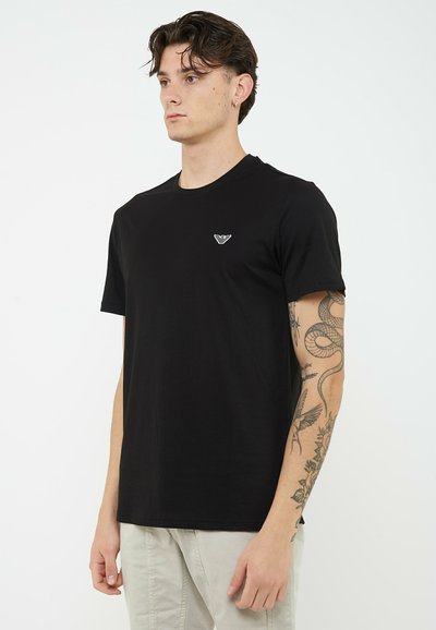 Joven con camiseta negra con un pequeño logo blanco y pantalones de color claro, mostrando un detallado tatuaje de serpiente y flores en su brazo.