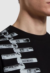 T-shirt in cotone nero con colletto rotondo, caratterizzata da un design grafico bianco e dal testo "Bikkembergs" sulla spalla sinistra.