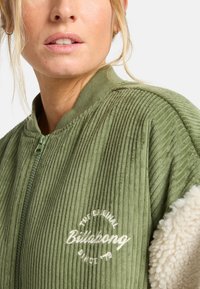 Kobieta ubrana w zieloną kurtkę z prążkowanego materiału, z kremowymi rękawami z sherpa, zapinaną na zamek z białym logo "Billabong The Original Since '78" na klatce piersiowej.