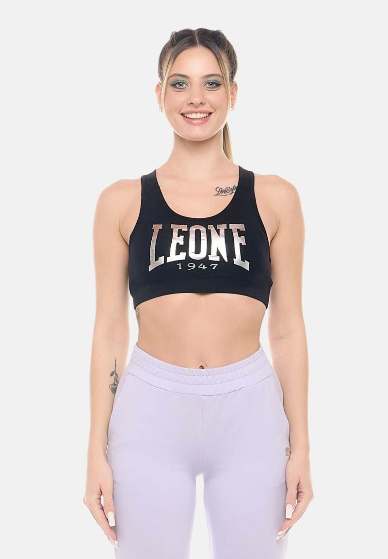 Jeune femme souriante, portant un soutien-gorge de sport noir avec le texte « LEONE 1947 » et un pantalon taille haute violet clair, debout sur un fond blanc.