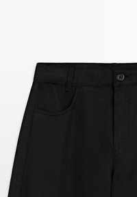 Sorte bukser i taljeområdet med bæltestropper, knaplukning og forlommer mod en ensartet lys baggrund.