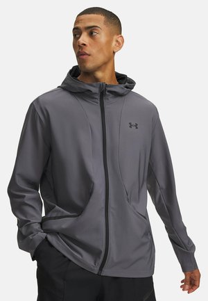 Man draagt een grijze zip-up capuchonjas met zwarte broek, kijkt opzij met één hand in de zak en de andere ontspannen.