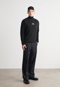 Black turtleneck sweater with logo detail, paired with dark, wide-leg trousers. Black cap and black shoes, minimalistic design and colors.