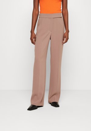 Pantalons larges couleur marron clair avec une texture lisse, présentant un devant plat et une fermeture éclair dissimulée, associés à des chaussures plates noires.