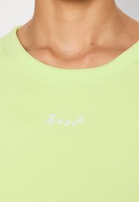 Gros plan sur le cou et le haut de la poitrine d'une personne portant un t-shirt côtelé vert clair avec le mot « "Bossy" » brodé en blanc.