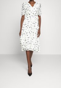 Robe enveloppante blanche à manches bouffantes courtes, présentant un motif à pois bleu et gris, une taille à nouer et une jupe fluide. Talons aiguilles noirs.