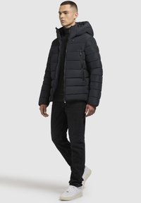 Mörk vadderad jacka med huva, med dragkedja fram, horisontell sömnad och sidofickor. Bärs över en svart tröja och jeans.