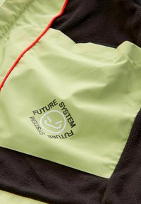 Hellgrüner Stoff mit schwarzer Fleece-Struktur, versehen mit einem Logo mit dem Schriftzug "FUTURE SYSTEM" und roten Akzentnähten entlang der Naht.