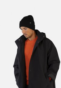 Gorro de punto negro, suéter naranja y abrigo impermeable negro. El abrigo tiene un cuello alto y cierres de presión, con detalles de costura visibles.