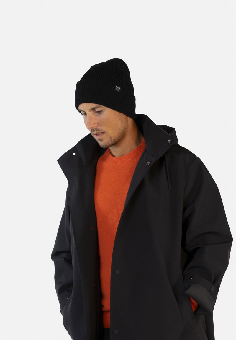 Gorro de punto negro, suéter naranja y abrigo impermeable negro. El abrigo tiene un cuello alto y cierres de presión, con detalles de costura visibles.