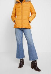 Skallig orange puffjacka med fake fur huva, dragkedja och två sidofickor, kombinerad med utställda blå denimjeans och mörka ankelstövlar.