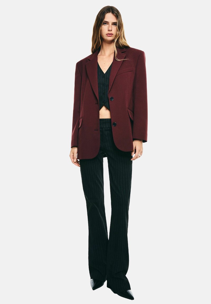 Ung kvinna bär en oversized bordeauxfärgad blazer, svart kortärmad knäppt topp, svart randig byxor och spetsiga svarta skor, står rakt.