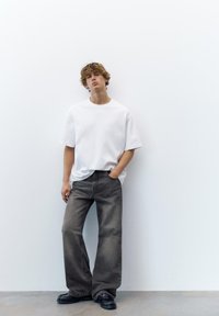 Junger Mann mit lockigem Haar, der ein locker sitzendes weißes T-Shirt, weite dunkelgraue Jeans und schwarze Schuhe trägt, steht vor einer weißen Wand.