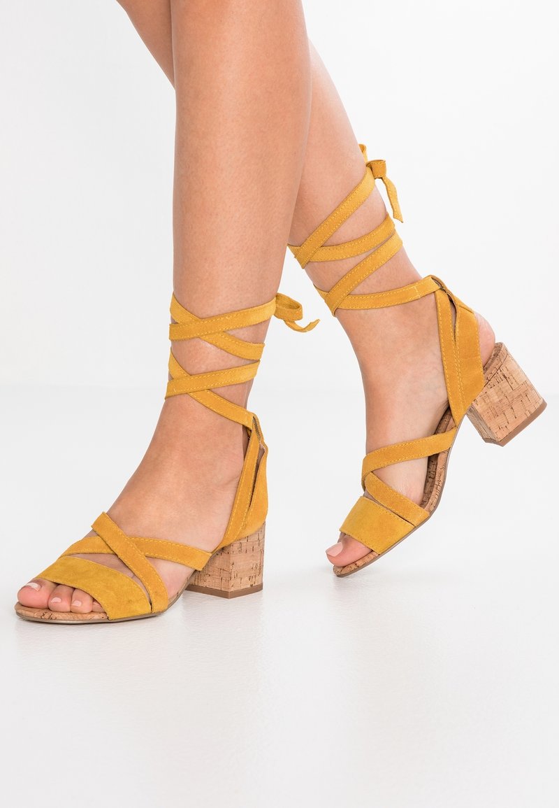 Pieds portant des sandales à talons carrés en daim jaune moutarde avec des lanières enveloppant la cheville sur un sol blanc.