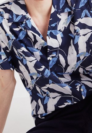 Homme portant une chemise à manches courtes boutonnée avec des motifs abstraits de feuilles bleues et blanches, penché légèrement vers l'avant.