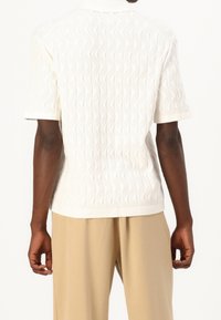 Wit gebreide korte mouw trui met een textuur van golvende patronen, geribde kraag en een ontspannen pasvorm, gecombineerd met lichtbeige broek.