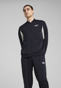 Marine tracksuit gemaakt van gladde, rekbare stof. Jas heeft een ritssluiting aan de voorkant, witte accenten en een logo. Broek is bijpassend met logo-detail.