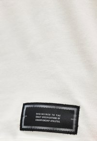 Μαύρη ορθογώνια υφασμάτινη ετικέτα σε ανοιχτόχρωμο ύφασμα με λευκό κείμενο: σχεδιασμένη σύμφωνα με τις ακριβείς προδιαγραφές αθλητών πρωταθλημάτων.