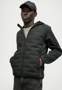 Zwarte pufferjas met ritssluiting en capuchon, met een gequiltde textuur en contrasterende mouwen, gecombineerd met een lichte trui en donkere jeans.