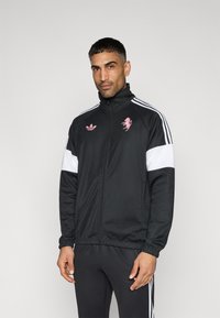 Μαύρο μπουφάν με φερμουάρ και λευκές ρίγες, με ροζ κεντητό λογότυπο μονόκερου και σήμανση Adidas στο μπροστινό μέρος. Λείο υλικό και εφαρμοστή σχεδίαση.