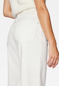 Valkoiset, korkeavyötäröiset housut suoralla lahkeella, joissa on takataskut, näkyvät ompeleet ja teksturoitu kangas.