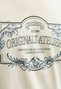 Σχεδιασμός περίτεχνου ετικέτας με το κείμενο «Originals Atelier», ανθισμένες διακοσμήσεις και φράσεις «Worldwide», «Est. 1990» και «Designed in Copenhagen.»