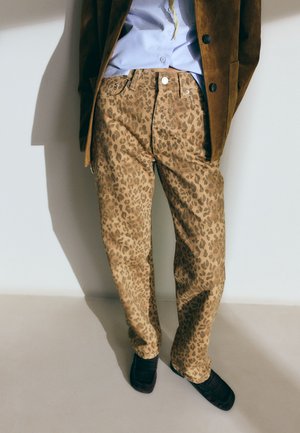 Henkilö, jolla on päällään leopardi-kuosiset housut, vaaleansininen paita, ruskea mokkatakki ja mustat kengät, seisomassa yksinkertaista vaaleaa taustaa vasten.