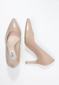 Une paire de chaussures à talons hauts en cuir verni beige, l'une montrant la vue de dessus avec l'étiquette de la semelle intérieure, l'autre en vue latérale avec un bout pointu et un talon aiguille.