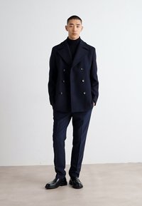 Két gombos sötétkék gyapjú kabát nagy fekete gombokkal, sötétkék slim-fit nadrággal és fekete bőrcipővel párosítva. Egyszerű, strukturált dizájn.