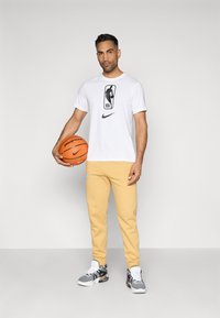 Άνδρας με λευκή μπλούζα NBA και κίτρινες φόρμες κρατώντας μια πορτοκαλί μπάλα μπάσκετ, που στέκεται μπροστά από ένα απλό ανοιχτό γκρι φόντο.