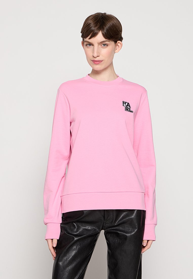 Hellrosa Sweatshirt aus weichem Stoff, mit langen Ärmeln und Rundhalsausschnitt. Die Vorderseite hat ein kleines schwarzes Logo-Detail. Getragen mit schwarzen Lederhosen.