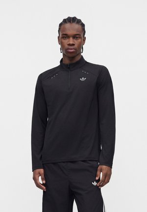 Junger Mann mit geflochtenem Haar, der einen schwarzen Adidas Viertel-Reißverschluss-Pullover und passende schwarze Adidas Trainingshose trägt.
