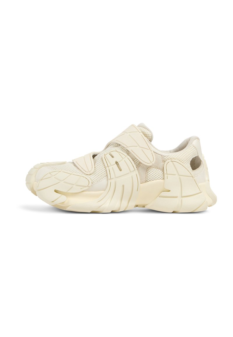 Beige atletiska skor med en texturerad mesh-övre del, kardborrebandsremmar och en formad gummisula med distinkta mönsterdetaljer.