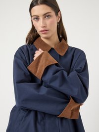 Νεαρή γυναίκα με μακριά καστανά μαλλιά, φορώντας σκούρο μπλε παλτό με γιακά και μανσέτες από καφέ κορντουρό ύφασμα, με σταυρωμένα τα χέρια, σε απλό φωτεινό φόντο.