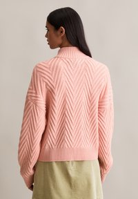 Femme aux longs cheveux noirs portant un pull en tricot texturé rose et une jupe en velours côtelé beige, de dos contre un fond uni.