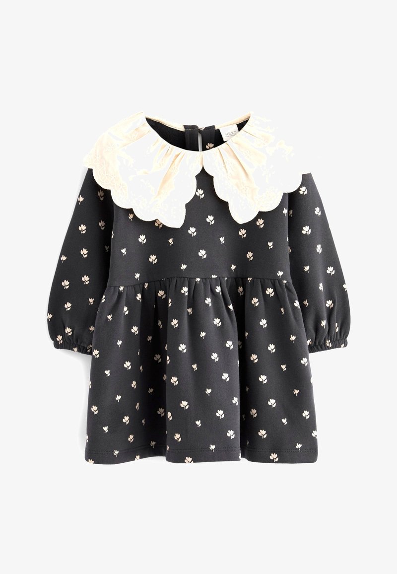 Vestido negro con pequeño estampado de flores blancas, que presenta un gran cuello crudo con forma de ondas, mangas largas y una falda acampanada.