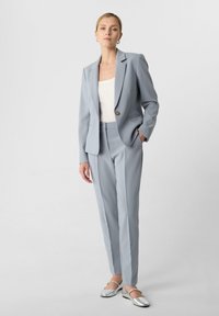 Lichtblauwe op maat gemaakte blazer en bijpassende broek van gladde stof, aangevuld met een witte top en zilveren platte schoenen.