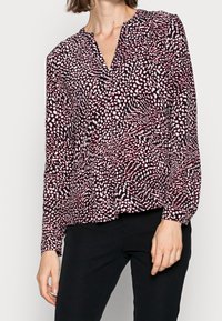 Femme portant un chemisier noir à manches longues avec un motif abstrait de points blancs et rouges et un pantalon noir, vue du cou aux hanches.