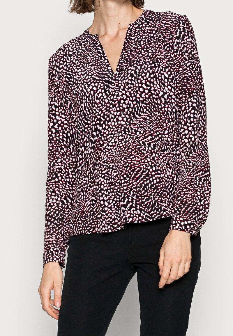 Femme portant un chemisier noir à manches longues avec un motif abstrait de points blancs et rouges et un pantalon noir, vue du cou aux hanches.