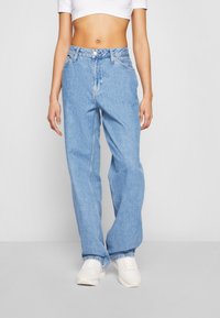 Kvinna i ljusblå löst sittande jeans, vit kort topp och vita sneakers, som står mot en enhetlig ljusgrå bakgrund.