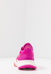 Jasně růžová sportovní bota s bílou mezipodešví a korálovými akcenty, zobrazená ze zadní strany na bílé ploše a pozadí.