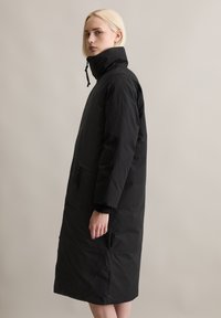 Schwarzer langer Mantel aus strukturiertem Stoff, mit hohem Kragen, seitlichen Taschen und geradem Schnitt, der unter das Knie fällt.