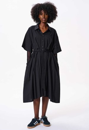Robe chemise oversize en coton noir avec col, devant à boutons, poches latérales et ceinture en tissu, portée avec des baskets noires.